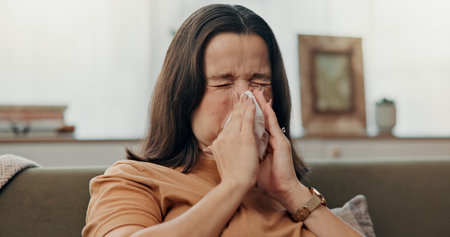 Home, sneeze and woman on sofa, tissue and allergies with season cold in living room. Person, apartment and lady on couch, flu and sick with illness, disease and burnout with cough, rest and sinusの写真素材
