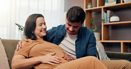 Pregnant woman, man and touch stomach on sofa, smile or laugh for baby kick, connection and love in house. Couple, parents and hug with care, support and happy with pregnancy for growth with bellyの写真素材