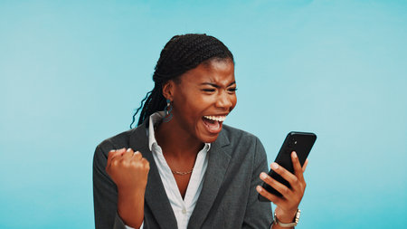 Excited, phone and black woman with celebration in studio for winning notification, success news or online reward. Mockup space, person and victory fist on blue background for achievement or giveawayの写真素材