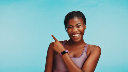 Black woman, fitness and portrait in studio with pointing, sports announcement or happy with space. Personal trainer, smile or showing gesture on blue background for gym sign up promo or membershipの写真素材