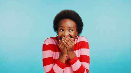 Secret, woman and laughing for gossip in studio with funny story, confidential information or surprise news. Mockup space, scandal or black person with mouth cover for private joke on blue backgroundの写真素材