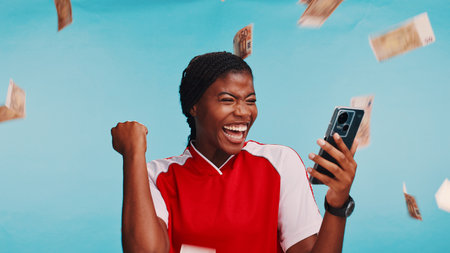 Woman, phone and sports betting in studio with money rain, excited and celebration by blue background. African girl, cash and happy with winning, competition and success with gambling on mobile appの写真素材