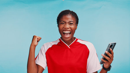 Black woman, phone and success in studio for sports, excited and selection for soccer team by blue background. Girl, athlete and smartphone in portrait, good news and celebration for draft in USAの写真素材