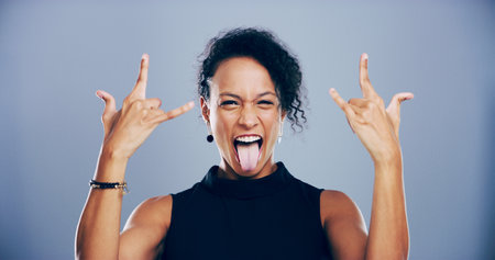 Crazy, tongue out and woman with rock hand gesture, studio or portrait for heavy metal on gray background. Culture, edgy female rocker or person with devil horn sign, funky emoji or punk symbolの写真素材