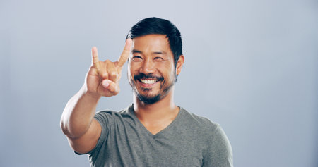 Rock, emoji and portrait of Asian man in studio with hand gesture for crazy, heavy metal and edgy attitude. Mockup space, happy and person with punk sign, symbol and devil horns on gray backgroundの写真素材