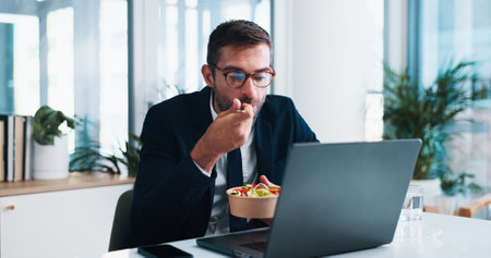 Business, man and eating with laptop in office for working lunch, reading economy news or report deadline. Financial analyst, employee or healthy salad bowl with tech for nutrition or market researchの写真素材