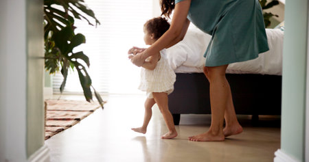 Mother, child and learning to walk in home with holding hands, first steps or support with milestone. Happy family, baby and woman with teaching balance in bedroom with mobility or growth developmentの写真素材