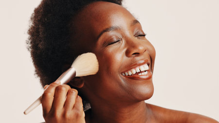 Brush, powder and black woman in studio with face, beauty or cosmetics for getting ready. Happy, cosmetology and African female person with blush for facial color or glow by white background.の写真素材