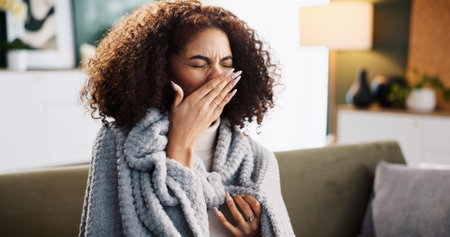 Sneeze, sick and tired with woman on sofa for medical recovery, illness and flu season. Disease, fatigue and fever with female person and blanket in living room at home for virus outbreak and coldの写真素材