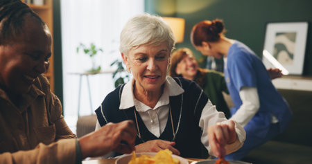 Senior women and retirement with eating in home with conversation, bonding and wellness with snacks. Elderly, people and friends in living room with chips for relax, support and happy at dinner tableの写真素材