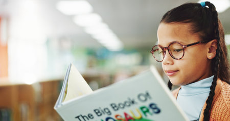 Girl, child and reading book at library for education, learning or concentration with dinosaur story. School kid, glasses and scholarship with language, knowledge and study for development at academyの写真素材