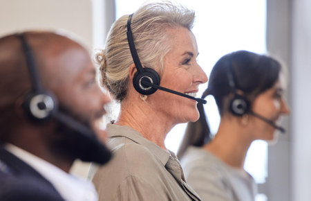 Call center, headset and mature woman in office for customer service, consulting or telemarketing. Agent, contact us and female with colleagues for communication, assistance or tech supportの写真素材