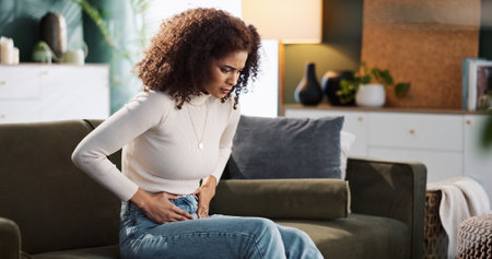 Couch, woman and hands on stomach for pain, menstrual cycle or endometriosis in lounge at home. House, female person or sick with abdomen ache for constipation, gastro crisis or period cramps on sofaの写真素材