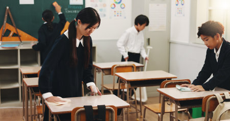Students, girl and cleaning desk at school for hygiene, learning responsibility and discipline in classroom. Japanese academy, people and wipe surface for disinfection, germs prevention and teamworkの写真素材