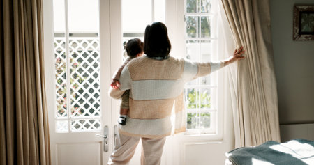 Mother, kid and open curtain in home for start day, morning routine and fresh air. Family, back and woman with baby by bedroom window for getting ready, waking up and checking weather in apartmentの写真素材
