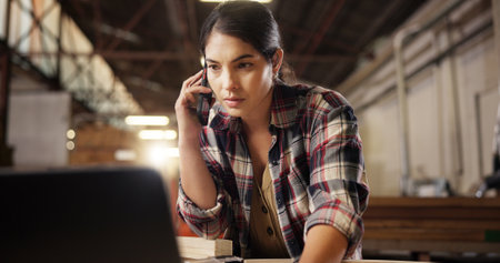 Phone call, laptop and woman in carpentry workshop for furniture, manufacturing and lumber renovation. Construction, factory and carpenter on smartphone for online orders, inventory and timber stockの写真素材
