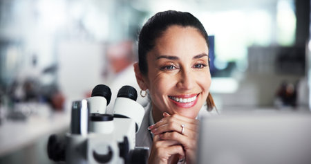 Computer, scientist and smile with woman with microscope for healthcare, medical research or test results. Tech, thinking or biologist breakthrough in clinical trial for pharmaceutical developmentの写真素材