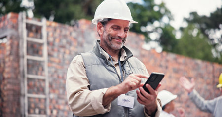 Construction site, smile and man with smartphone, typing and communication with property. Mature person, employee and inspector with cellphone, connection and building development with renovationの写真素材