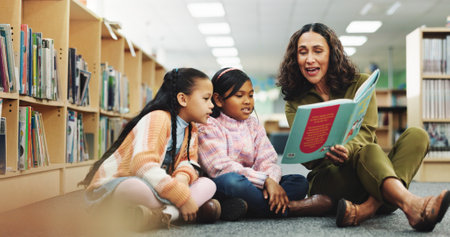 Girl, teacher and book with reading at library for development, learning or study with guide. School kids, storytelling and woman with help on floor, education and language for knowledge at academyの写真素材