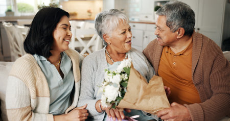 Birthday, flowers or gift with family on sofa in living room of home together for bonding or celebration. Bouquet, love or present with senior parents and daughter in apartment for event or milestoneの写真素材