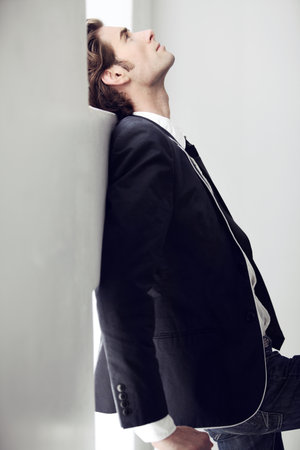 Depressed, sad and young man with fashion for formal style by a white wall in a modern home. Profile, classy style and male person from Canada in elegant suit with mental health issue in a space.の写真素材