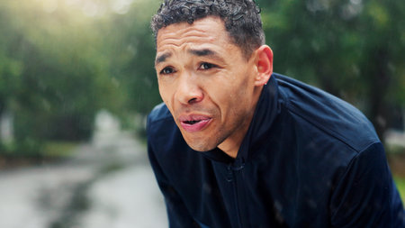 Fitness, man and tired with breathing at park for running burnout, workout recovery and cardio challenge. Athlete, person and rest in rain with exercise break, exhausted and overworked with trainingの写真素材