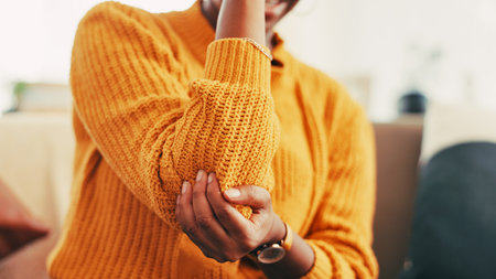 Elbow, injury and pain for person, home and joint with inflammation in living room, cramp and hurt. Hands, accident and woman with muscle tension in house, healthcare and symptoms of arthritisの写真素材