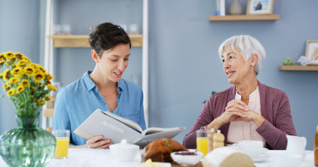 Senior woman, caregiver and reading book in home, breakfast or smile in retirement in dining room. People, connection and literature in morning for care, storytelling and support with novel in houseの写真素材