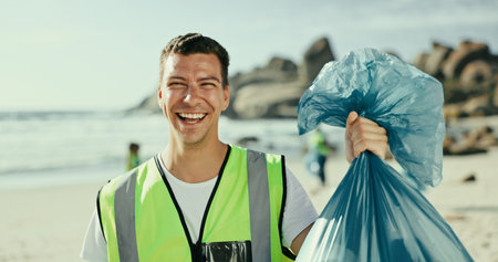 Man, smile and plastic bag for cleaning beach in portrait, outdoor or excited on earth day with care for environment. Person, volunteer and happy by sea for accountability with trash, garbage or dirtの写真素材