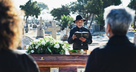 Bible, coffin and priest by graveyard with family for spiritual farewell, grief or loss. Religion, casket and pastor with people for memorial service at burial with holy book for blessing or goodbye.の写真素材
