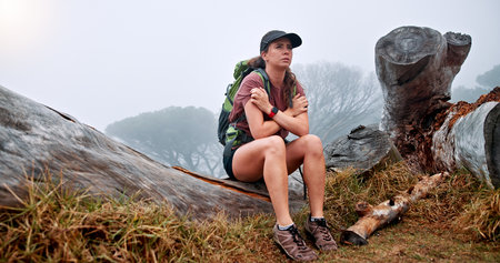 Girl, hiking and getting cold on mountain in winter, thinking and fatigue with weather change, fog and tourism. Person, woman and trekking on tree trunk, exhausted and tired on adventure in Argentinaの写真素材