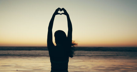 Sunset, beach and woman with heart hands for travel, vacation or getaway with calm in evening. Ocean, silhouette and back of female person on seaside weekend trip with love hand gesture or emoji.の写真素材
