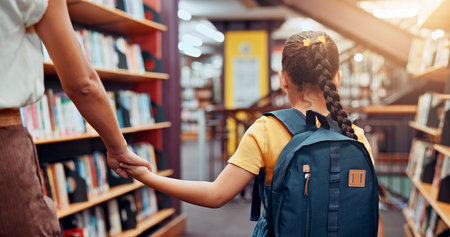 Education, tutor and child in library, holding hands and walking with student, bookshelf and back. Knowledge, growth and development of kid with books, learning and teacher with girl in schoolの写真素材