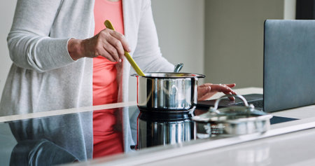 Woman, hands and cooking on stove with laptop, recipe and reading a blog about food. Kitchen, pot or person mixing with wood spoon, scroll online or learning to prepare meal or dinner with nutritionの写真素材