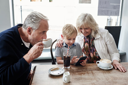 Family, kid and cafe with phone, games and click with smile for soda, relax and watch video. Grandparents, typing and child with tea cup, coffee shop and smartphone with reading, press and cartoonの写真素材