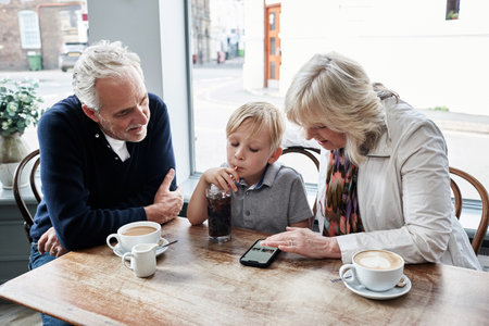 Man, woman and boy with phone in coffee shop, drink and soda with click, relax and watch video. Grandparents, family and child with tea cup, online and smartphone with reading, press and cartoonの写真素材