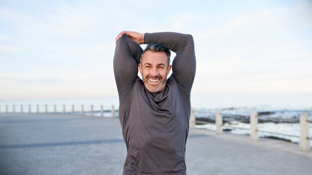 Happy, mature man and stretching by beach to start, getting ready and muscle preparation outdoor. Athlete, arms warm up and smile at promenade for cardio, wellness challenge and exercise performanceの写真素材