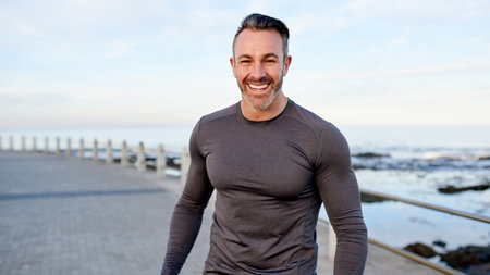 Mature man, runner and happy on promenade by ocean, break or pride with portrait for workout in morning. Person, training and fitness by sea with confidence, smile or outdoor for exercise in Spainの写真素材