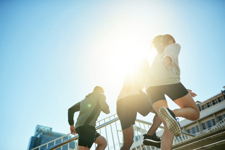 Fitness, group and running in city for health, body wellness or friends training in low angle. Team, cardio and people exercise outdoor on stairs for workout, sport or race with energy on sky spaceの写真素材