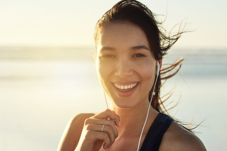 Happy woman, portrait and earphones on beach for fitness, self affirmation audio and windy at sunset. Athlete, smile and tech at Colombia ocean for workout music, local radio and motivation podcastの写真素材