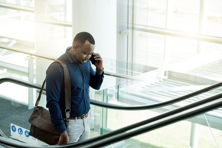 Phone call, office and business man on escalator for commute, travel and arrival in lobby. Modern building, conference and African person on electric stairs with smartphone for contact and networkingの写真素材