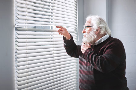 Curious senior man looking through window blinds while thinking, planning and wishing to escape old age home. Stressed, anxious or lonely elderly pensioner feeling trapped during covid lockdown alone
