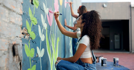 Girl, teamwork or artists painting on wall for mural, drawing flowers or community service at building. People, creative or women with brush for floral decoration, renovation or project for designの写真素材