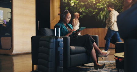Business woman, headphones and tablet in lobby for planning, research or travel for seminar. Chair, music or streaming with happy flight passenger in hotel with luggage or suitcase for job tripの写真素材