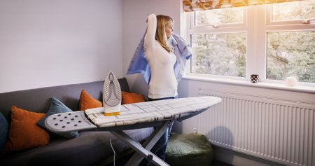 Iron, board and woman with shirt in morning, press cleaning or dressing for work as a cleaner in apartment. Ironing, laundry and person in living room with uniform, blouse or clean clothes in homeの写真素材