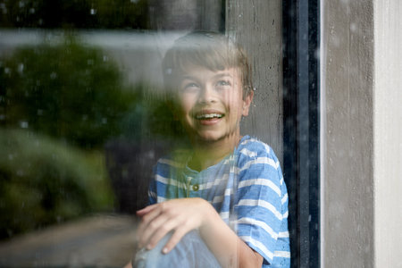 Happy, child and rain by window in home for sensory experience, imagination and soothing sound. Boy...