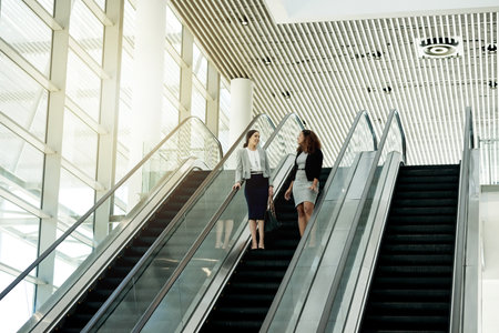 Office, escalator and business women in conversation in lobby for meeting, discussion and networking. Professional, corporate and people on stairs at conference, convention and seminar in buildingの写真素材