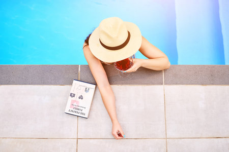 Top view, woman and drink at swimming pool for relax, wellness and freedom for luxury holiday. Water, book and female person at hotel for summer, travel and cocktail for vacation in Hamptonsの写真素材