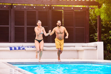 Man, woman and holding hands at hotel by swimming pool for travel, jump on holiday for wellness. Couple, running and together with support for vacation in Bali, dive and bonding for relationshipの写真素材