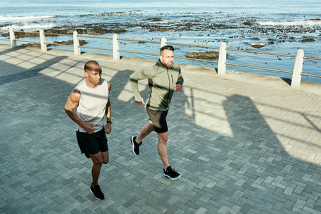 Men, running or fitness by beach promenade in exercise, workout or wellness with sports coach. Friends, team or personal trainer with athlete for marathon, race or competition training accountabilityの写真素材